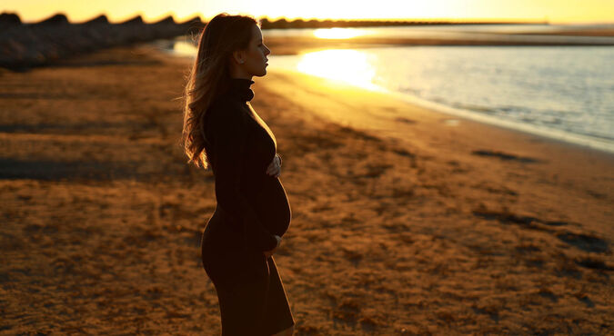 Mulher grávida em perfil segurando a barriga ao pôr do sol na praia
