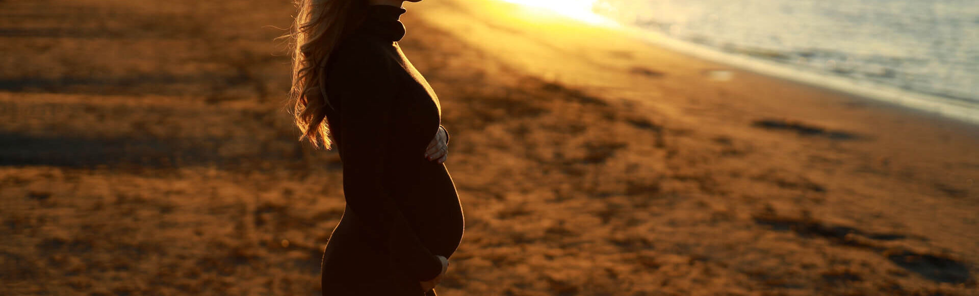 Mulher grávida em perfil segurando a barriga ao pôr do sol na praia
