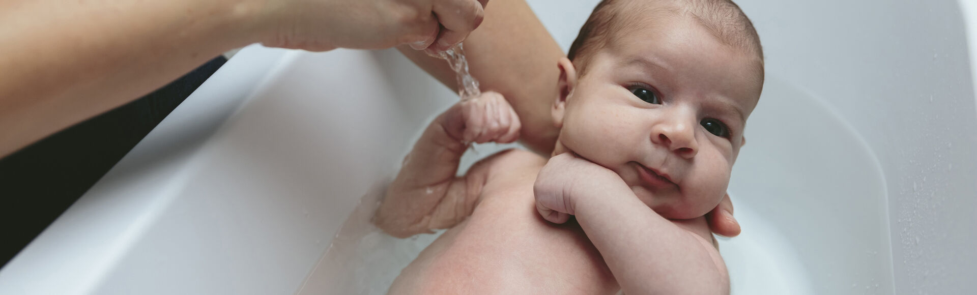 Bebê tomando banho na banheira com um adulto segurando-o por trás do pescoço