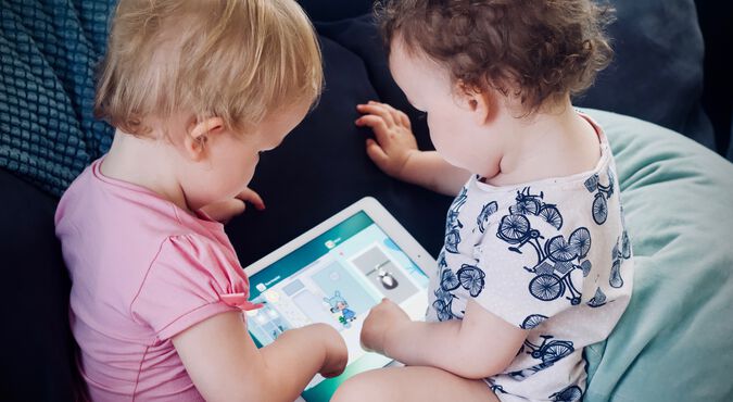 Criança e bebê à frente de um tablet com a supervisão da mamãe