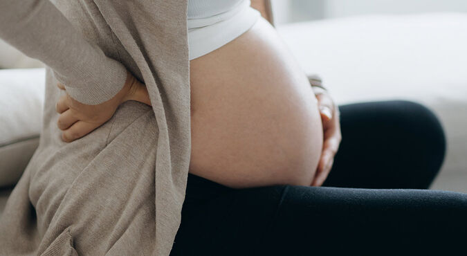 Gestante branca sentada com pernas cruzadas em frente ao corpo e mãos apoiadas na coluna lombar