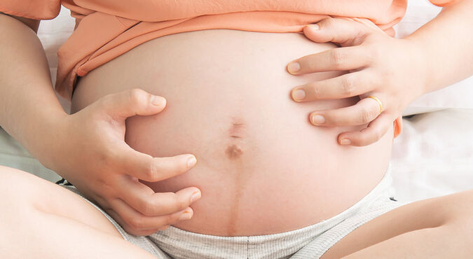 Gestante sentada com a barriga descoberta e as mãos ao redor do umbigo.