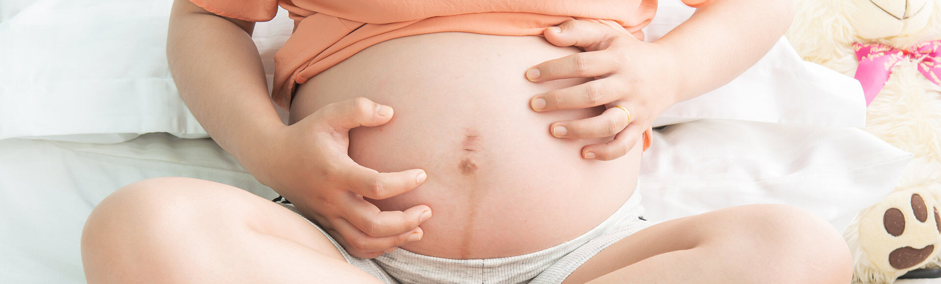 Gestante sentada com a barriga descoberta e as mãos ao redor do umbigo.