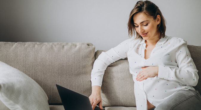 Gestante sentada com a mão na barriga enquanto usa um computador notebook