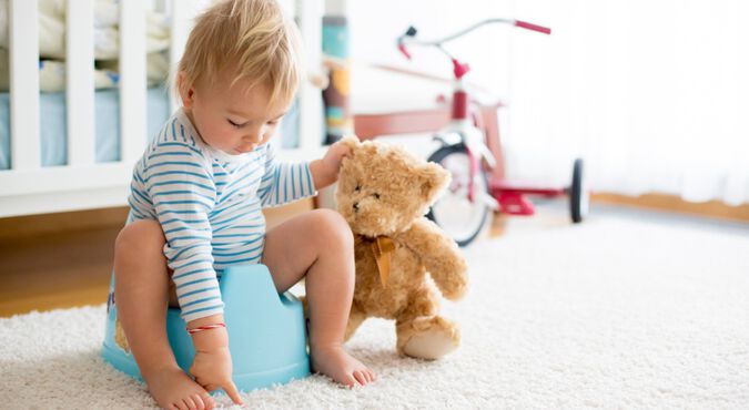 Criança sentada em um penico azul brincando com um ursinho de pelúcia em um quarto infantil