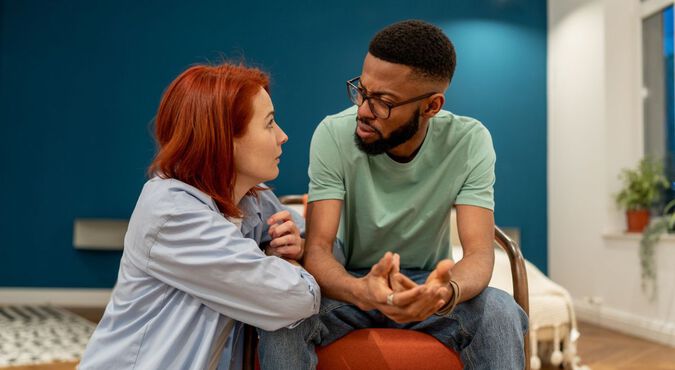 casal sentado em uma sala, aparentemente tendo uma conversa séria. A mulher, com cabelos ruivos, está inclinada para frente, enquanto o homem, com barba e óculos, está gesticulando com as mãos.