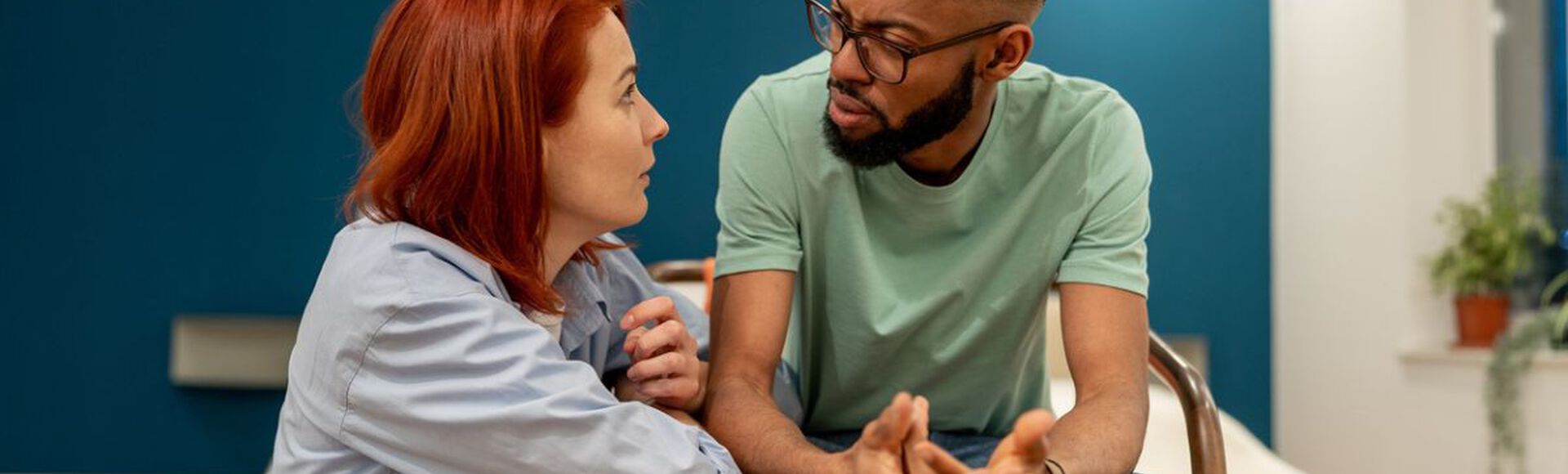 casal sentado em uma sala, aparentemente tendo uma conversa séria. A mulher, com cabelos ruivos, está inclinada para frente, enquanto o homem, com barba e óculos, está gesticulando com as mãos.