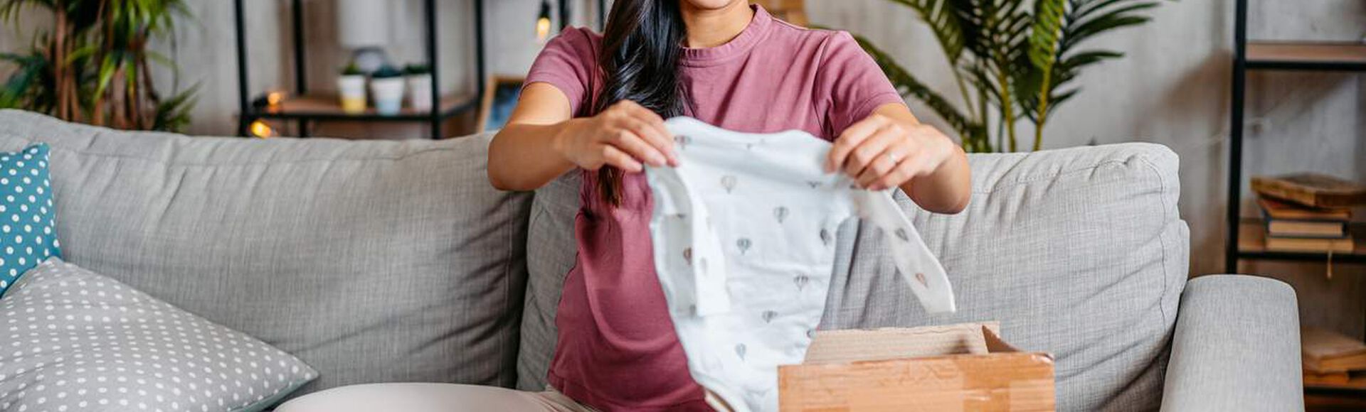 Mulher gestante abrindo um recebidinho: de presente, um pequeno macacão azulado.
