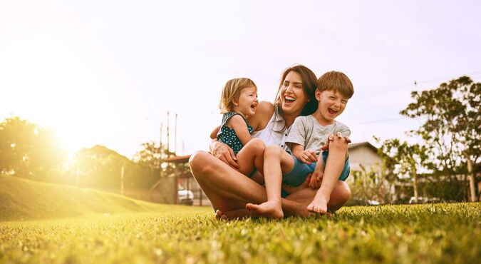 Mãe brincando com seus dois filhos pequenos em um gramado ao entardecer