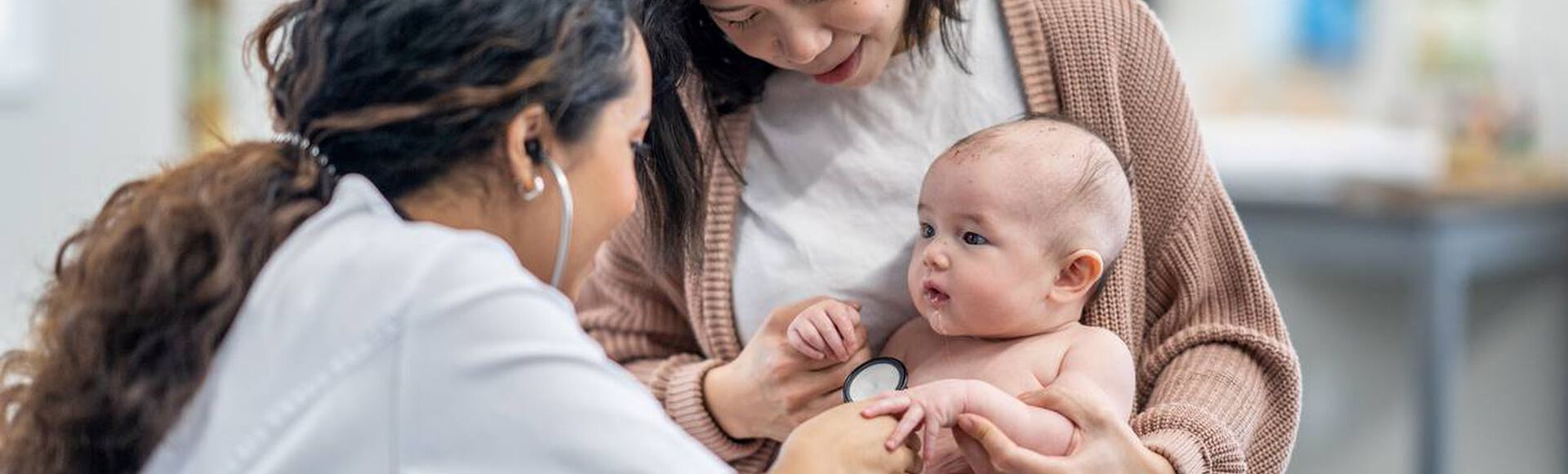 Médica examinando bebê no colo da mãe em uma consulta pediátrica.