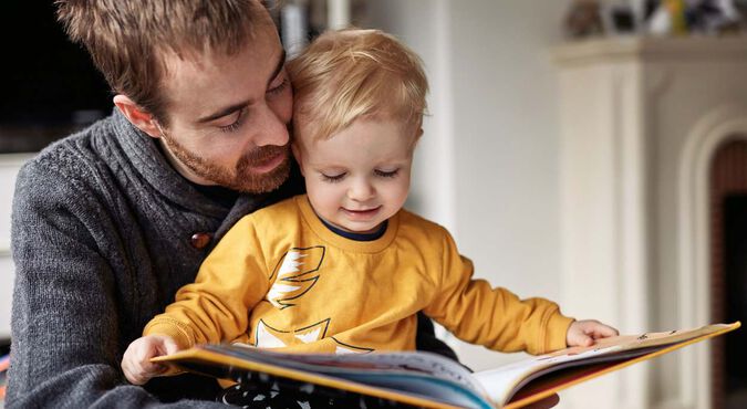 Pai segurando seu bebê no colo, com um livro aberto, lendo para ele