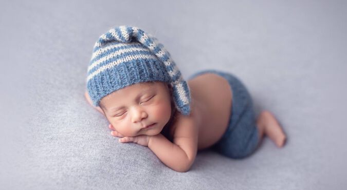 bebê dormindo tranquilamente em uma superfície macia. O bebê está usando uma touca listrada azul e branca e uma fralda ou calça azul combinando.