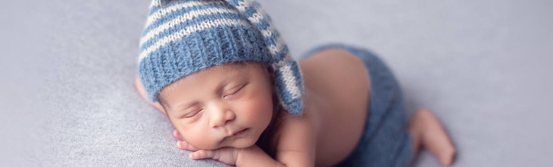 bebê dormindo tranquilamente em uma superfície macia. O bebê está usando uma touca listrada azul e branca e uma fralda ou calça azul combinando.