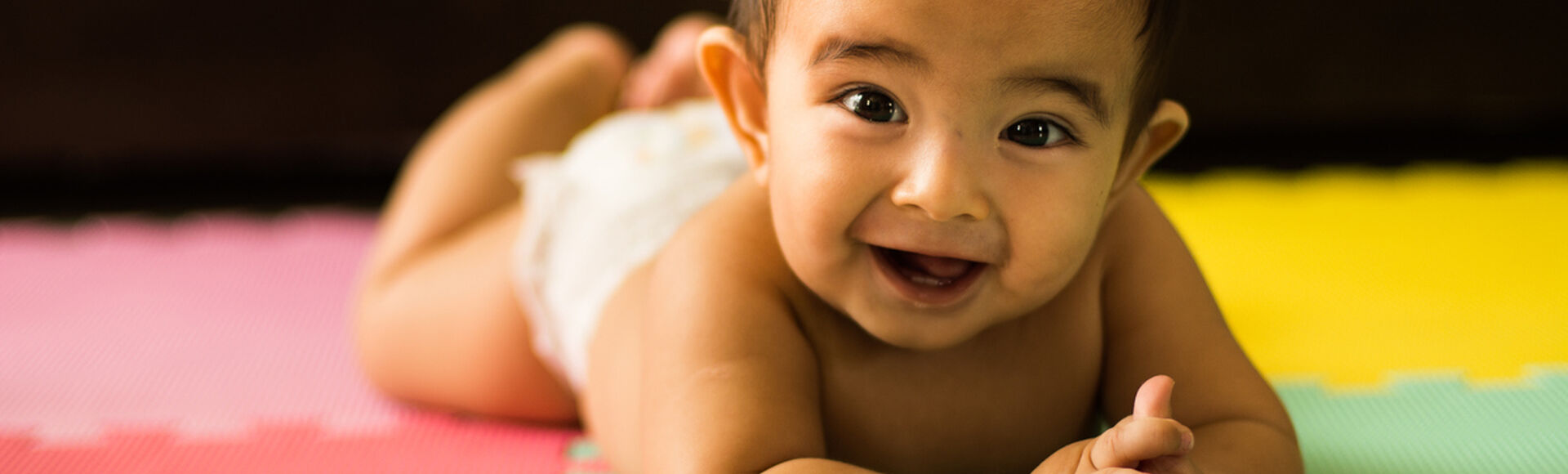 Bebê de bruços sobre tapete colorido de EVA, sorrindo e com a cabecinha levantada, bem firme.