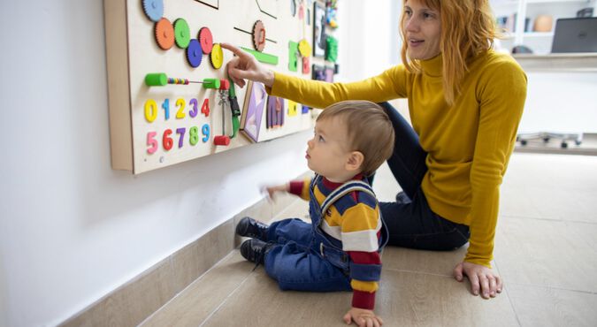 Mulher e bebê interagindo com painel de brincar. Eles são brancos, estão sentados no chão e usam roupa colorida.