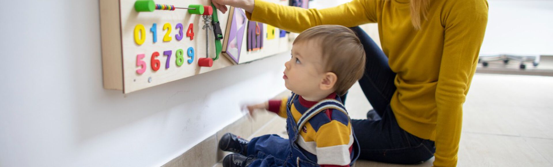 Mulher e bebê interagindo com painel de brincar. Eles são brancos, estão sentados no chão e usam roupa colorida.