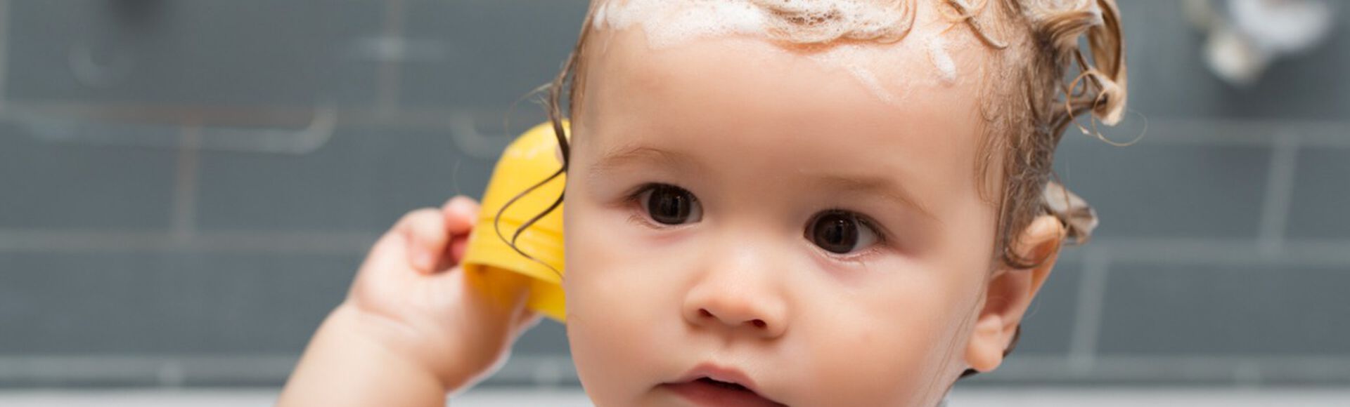 Bebê branco tomando banho de banheira com espuma de shampoo de bebê em seus cabelos castanhos. Ele aparece do ombro para cima