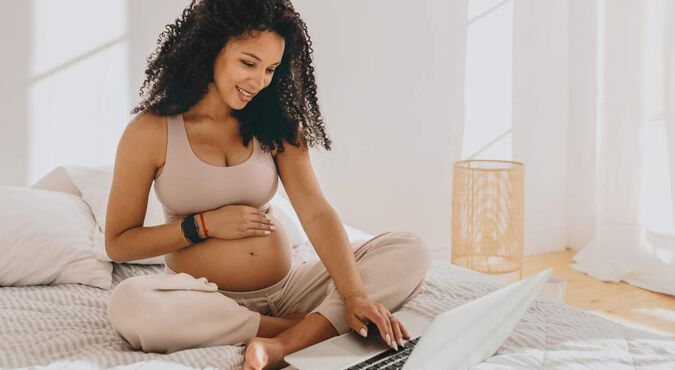Gestante sentada em posição de lótus na cama, com uma mão sobre a barriga e outra no laptop.