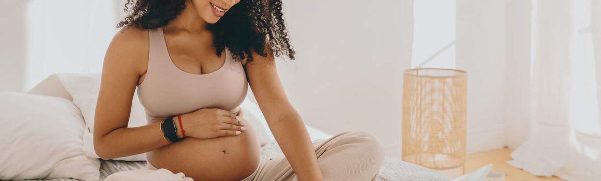Gestante sentada em posição de lótus na cama, com uma mão sobre a barriga e outra no laptop.
