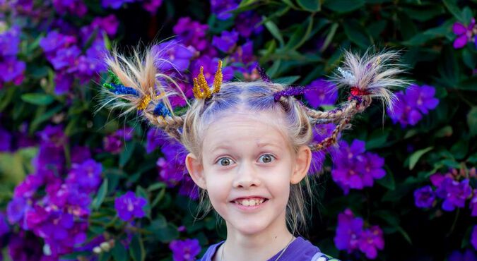 Garota feliz, com olhos arregalados e penteado inspirado nas flores roxas que estão no arbusto atrás dela.