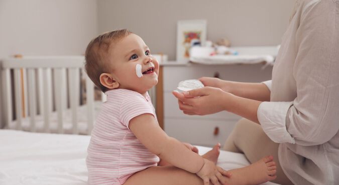 Bebê com body rosa claro listrado, sentada na cama com a mãe, enquanto tem seu rosto hidratado por creme apropriado