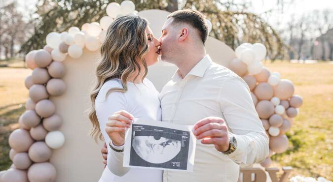 Casal se beijando enquanto segura um ultrassom, com decoração de balões neutros ao fundo