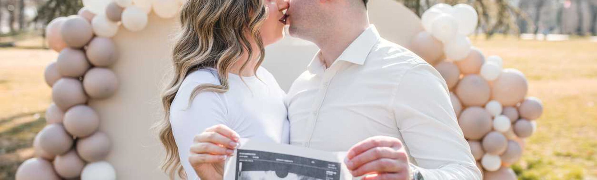 Casal se beijando enquanto segura um ultrassom, com decoração de balões neutros ao fundo