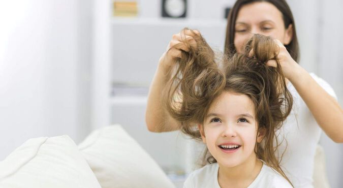 Mulher desembaraçando cabelo de menina que está sentada, olhando para cima. As duas vestem branco.