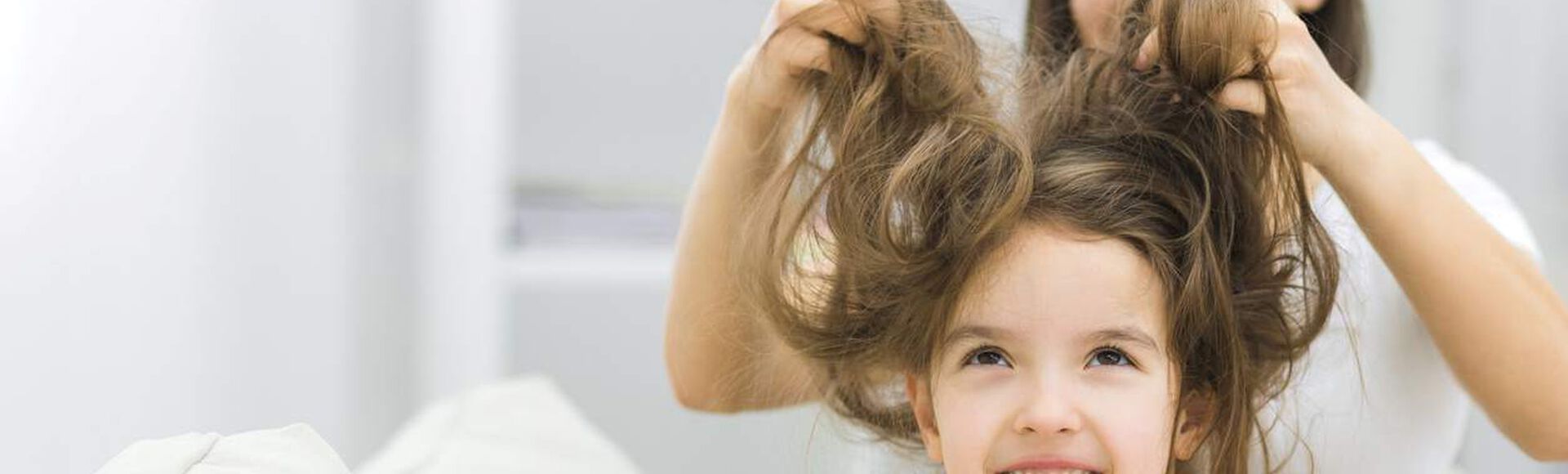 Mulher desembaraçando cabelo de menina que está sentada, olhando para cima. As duas vestem branco.