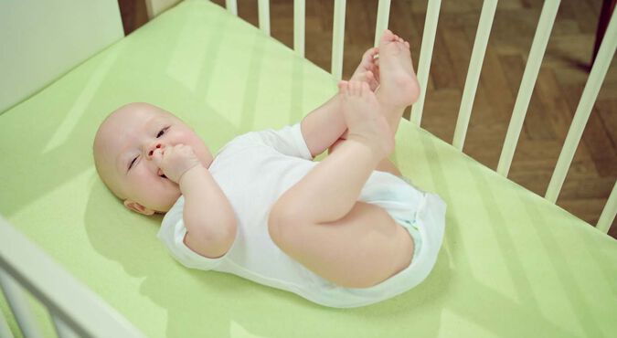 Bebê no bercinho, vestido de branco, com fralda noturna, brinca com uma mão na boca e outra nos pezinhos.
