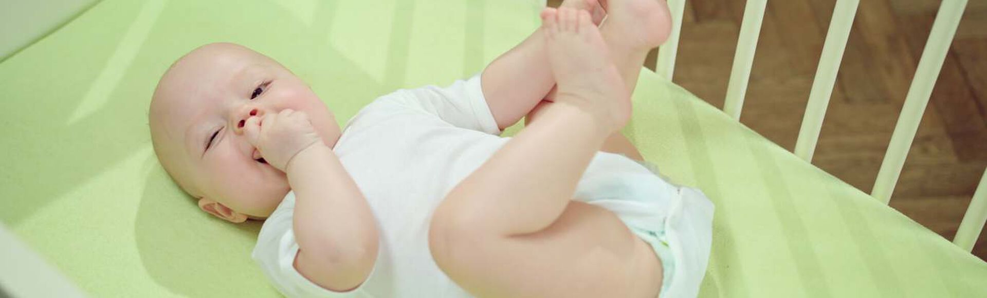 Bebê no bercinho, vestido de branco, com fralda noturna, brinca com uma mão na boca e outra nos pezinhos.