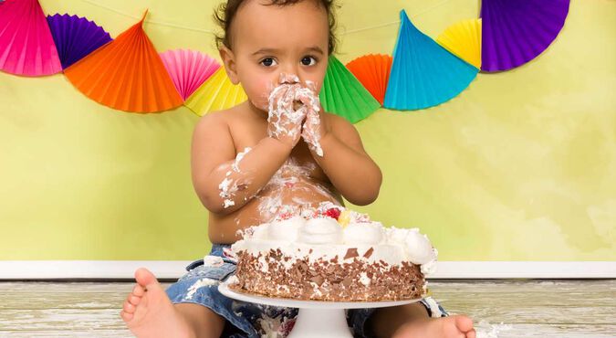 Bebê sentado, sem camisetinha, com chapéu de aniversário, está se divertindo ao provar um bolo direto com as mãos.