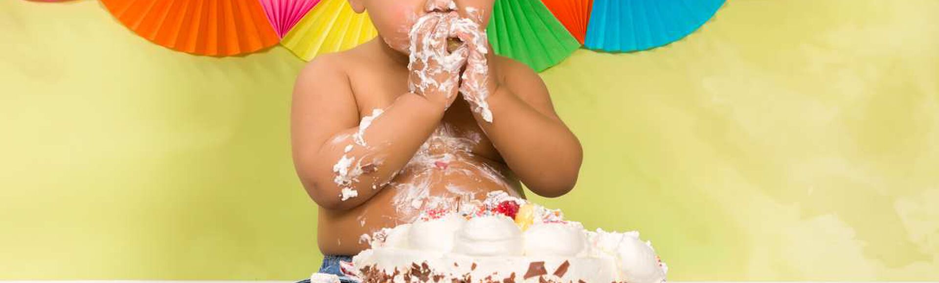Bebê sentado, sem camisetinha, com chapéu de aniversário, está se divertindo ao provar um bolo direto com as mãos.