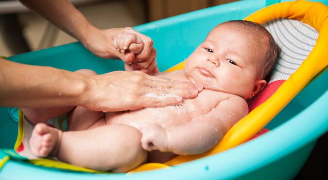 Bebê em banheira, sendo banhado por mãos cuidadosas de seu adulto responsável. Há espuma  sobre o corpinho.