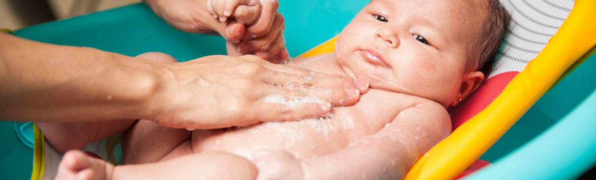 Bebê em banheira, sendo banhado por mãos cuidadosas de seu adulto responsável. Há espuma  sobre o corpinho.