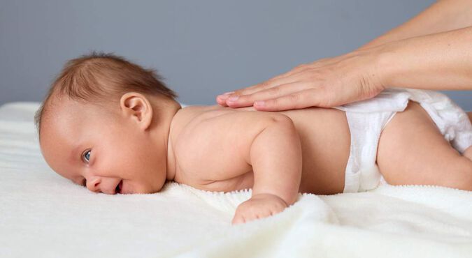 Bebê de bruços, sorrindo, sendo massageado nas costas por seu cuidador.