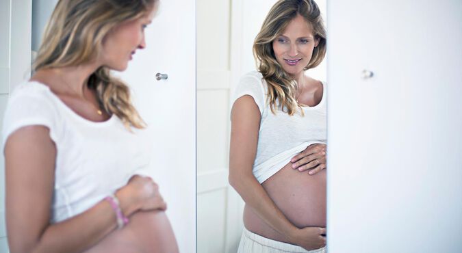 Mulher grávida admirando sua beleza em frente a um espelho, enquanto acaricia sua barriga gentilmente.