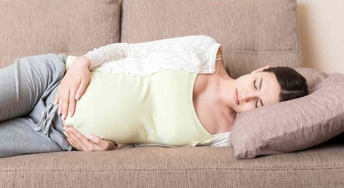 Mulher grávida ao celular, com uma mão no pé da barriga e fisionomia de dor