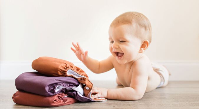 Bebê de bruços, sorrindo, dando tapinha em pilha de fraldas reutilizáveis.