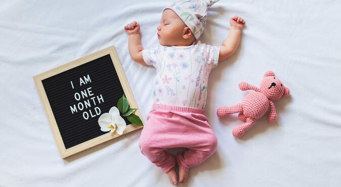 Bebê branca deitada em foto de mesversário. Ela veste rosa e branco, com plaquinha de 5 meses e ursinho ao lado