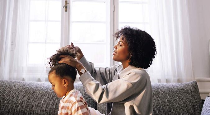 Mãe e filha na sala, sentadas no sofá, enquanto a mamãe penteia o cabelo de sua pequena.