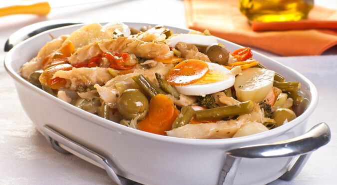 Travessa branca contendo bacalhau assado com azeitonas, ovos, cenouras e outros alimentos