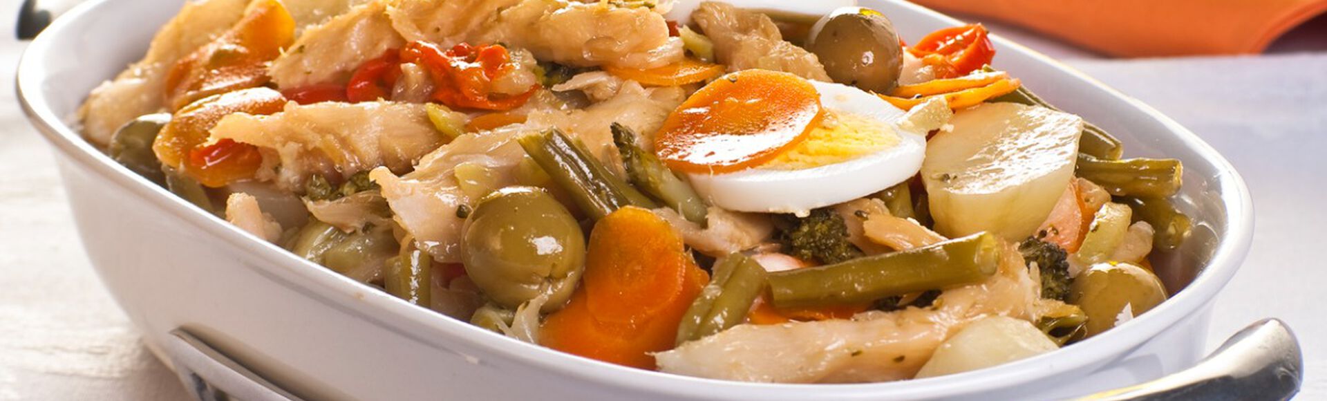 Travessa branca contendo bacalhau assado com azeitonas, ovos, cenouras e outros alimentos