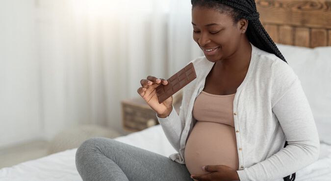 Mulher negra com tranças no cabelo segura barra de chocolate em cima da cama. Ela segura a barriga e olha para baixo sorrindo