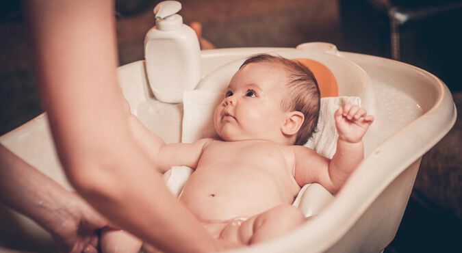 Bebê tomando banho em banheira com atenção voltada para o adulto.