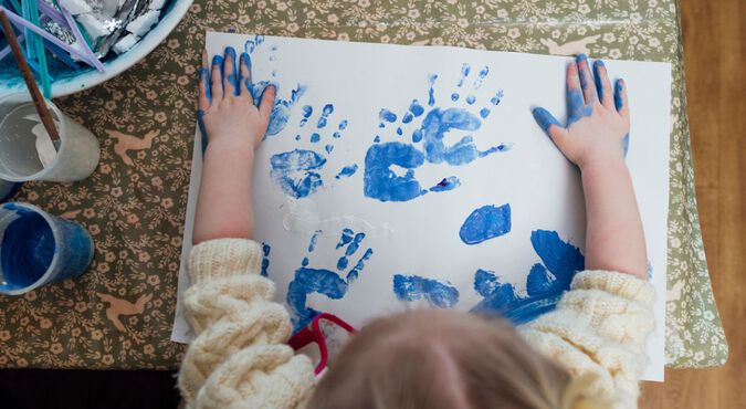 Criança fazendo arte com as mãos, usando tinta azul em uma folha de papel branco.