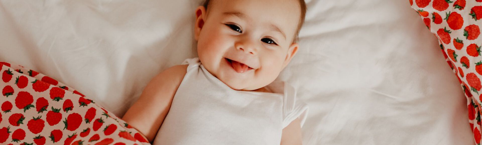 Bebê sorridente deitada e coberta com cobertor com estampa de morangos