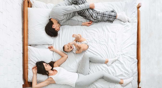 Bebê parece se divertir na cama, sorrindo e com perninhas para cima, enquanto seus pais cansados se encolhem nos cantos.
