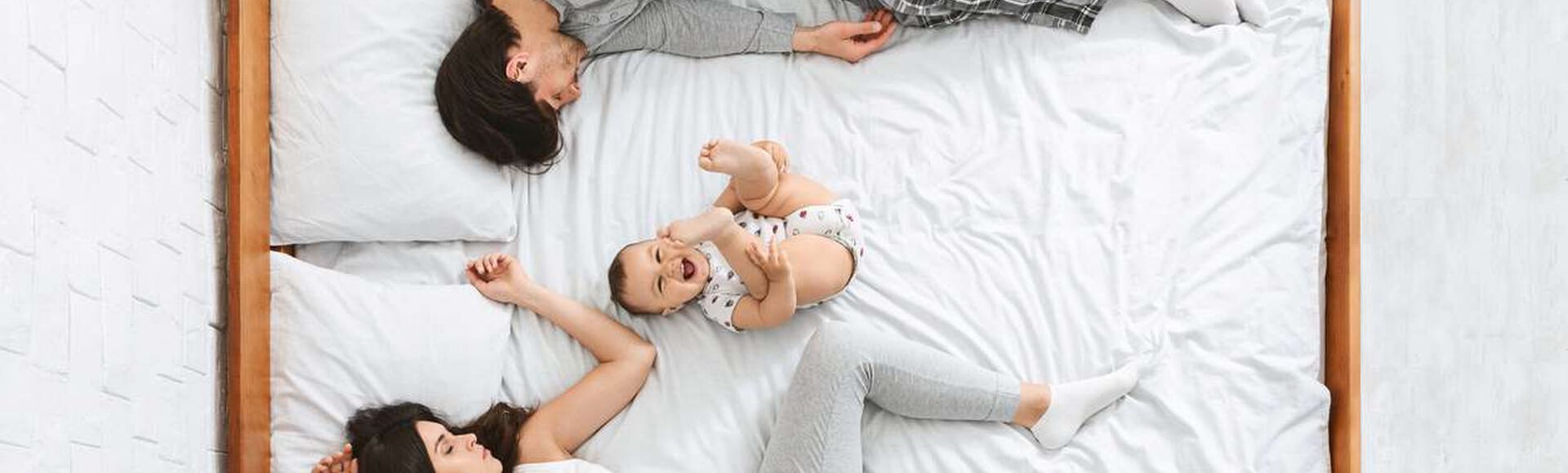 Bebê parece se divertir na cama, sorrindo e com perninhas para cima, enquanto seus pais cansados se encolhem nos cantos.