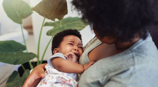 Mamãe consola bebê chorando na hora da bruxa. Não vemos o rosto da mãe que está olhando para o bebê no colo. Os dois são negros.